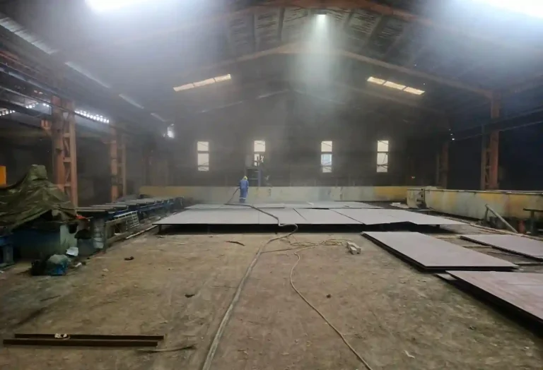 An interior view of a heavy fabrication workshop featuring large curved steel plates for storage tank fabrication services. Welding equipment and a giant industrial tank structure under construction are visible in the background, compliant with API and ASME standards.