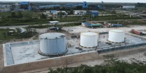 An aerial view of an industrial facility featuring three large white and gray storage tanks, set against a backdrop of a factory complex and a remote landscape. This serves as an example of a completed storage tank fabrication project.